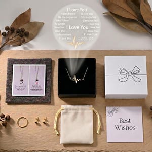 Op de afbeelding: Een assortiment sieraden en geschenkdozen op een getextureerd oppervlak. Inclusief een ketting in een zwarte doos, een ring, oorbellen, een klein zakje en een kaart met "Best Wishes". De afbeelding bevat ook de tekst "I Love You" in meerdere talen.