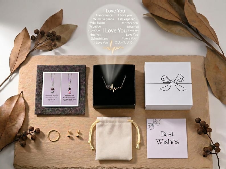 Op de afbeelding: Een assortiment sieraden en geschenkdozen op een getextureerd oppervlak. Inclusief een ketting in een zwarte doos, een ring, oorbellen, een klein zakje en een kaart met "Best Wishes". De afbeelding bevat ook de tekst "I Love You" in meerdere talen.