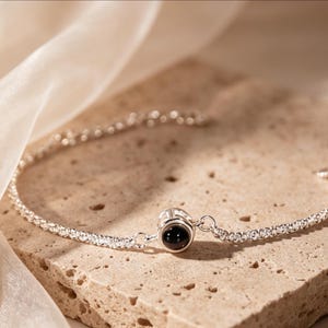 May include: A silver bracelet with a delicate chain design. The bracelet features a central charm with a black stone. The jewelry is displayed on a textured, light brown surface, with a soft, white fabric in the background.