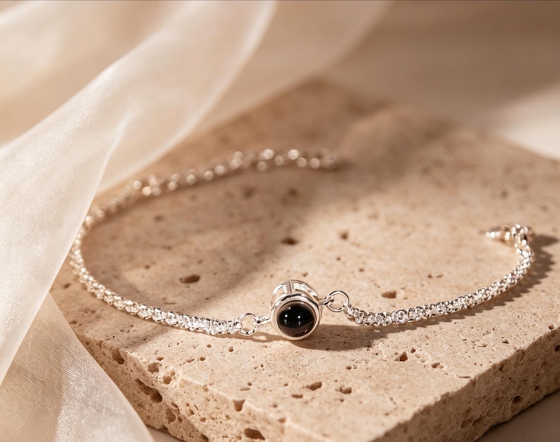 May include: A silver bracelet with a delicate chain design. The bracelet features a central charm with a black stone. The jewelry is displayed on a textured, light brown surface, with a soft, white fabric in the background.