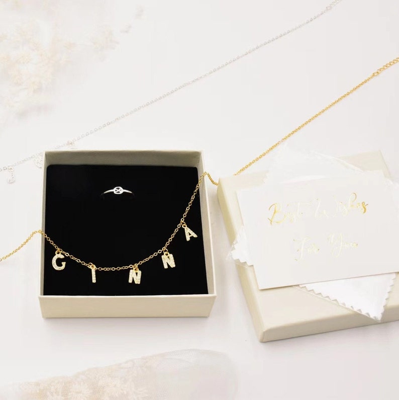 May include: A white box with a gold necklace with the name "GINA" spelled out in charms. The box also contains a small silver ring. The box is sitting on a white surface. The text "Best Wishes for You" is written on a white card inside the box.
