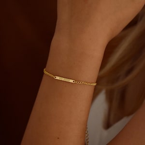 Könnte beinhalten: Ein goldfarbenes Armband mit einem rechteckigen Steg und Kettengliedern. Der Steg ist mit den römischen Ziffern VII XXIII graviert. Das Armband wird am Handgelenk getragen.