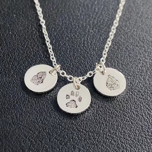 Op de afbeelding: Een zilveren ketting met drie ronde hangers. Elke hanger heeft een getextureerde afdruk, met &eacute;&eacute;n met een pootafdruk en de andere met bladeren. De ketting en hangers zijn gemaakt van glanzend zilver metaal.