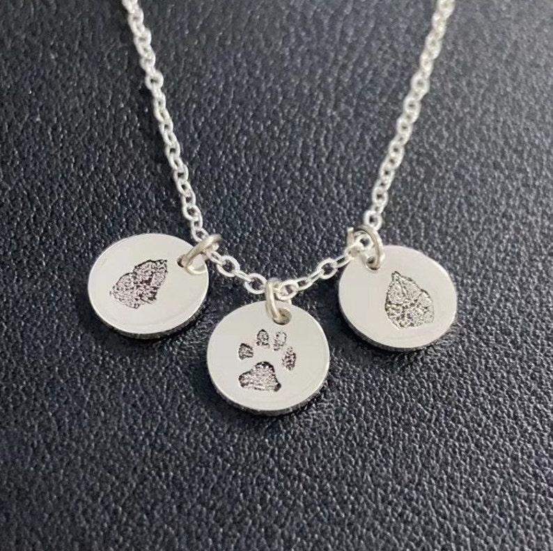 Op de afbeelding: Een zilveren ketting met drie ronde hangers. Elke hanger heeft een getextureerde afdruk, met &eacute;&eacute;n met een pootafdruk en de andere met bladeren. De ketting en hangers zijn gemaakt van glanzend zilver metaal.