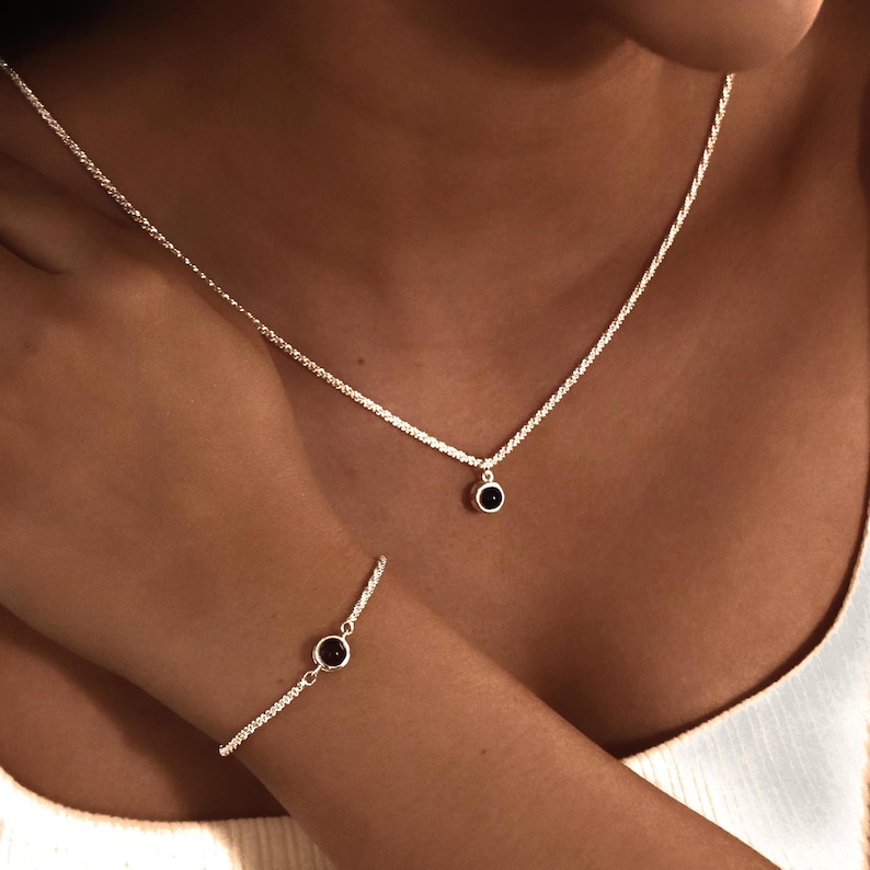 May include: A silver necklace and bracelet set. The necklace features a delicate chain with a small, round, black gemstone pendant. The matching bracelet has a similar design, with a single gemstone. The jewelry is displayed on a person.
