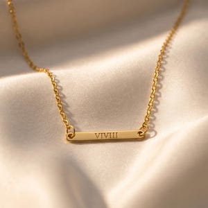 May include: A gold-colored necklace with a delicate chain and a rectangular pendant. The pendant features the engraved Roman numeral "VIVIII". The necklace rests on a soft, cream-colored fabric, creating a luxurious aesthetic.