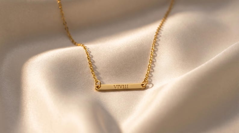 May include: A gold-colored necklace with a delicate chain and a rectangular pendant. The pendant features the engraved Roman numeral "VIVIII". The necklace rests on a soft, cream-colored fabric, creating a luxurious aesthetic.