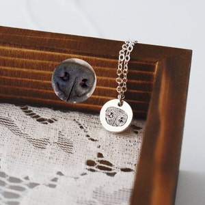 Op de afbeelding: Een zilveren hangerketting met een cirkelvormige bedel met een gedetailleerde afdruk van een hondenneus. De ketting hangt aan een fijne ketting. Een aparte afbeelding van de hondenneus staat op de achtergrond. De ketting wordt getoond op wit kant.