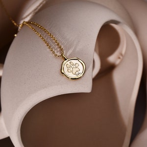 May include: A gold necklace with a small, round pendant featuring a paw print design. The pendant is on a light brown background.