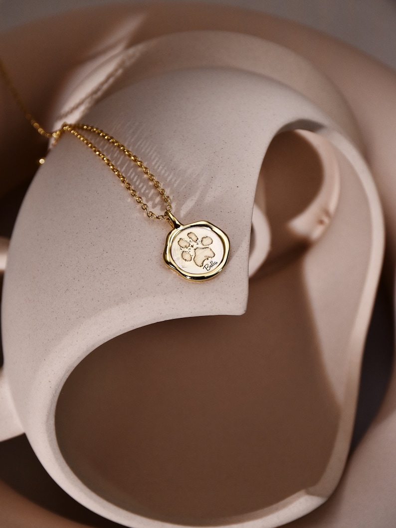 May include: A gold necklace with a small, round pendant featuring a paw print design. The pendant is on a light brown background.