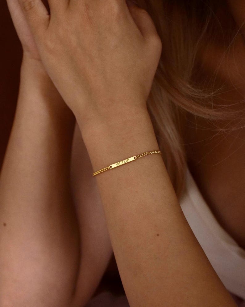 May include: A gold-toned chain bracelet featuring a central rectangular bar engraved with the Roman numerals VII XXIII. The bracelet consists of a delicate link chain that secures around the wrist, showcasing a polished metallic finish throughout.
