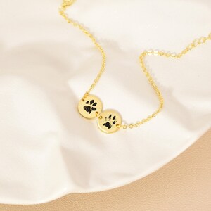 May include: A gold necklace with two circular pendants, each displaying a black paw print. The necklace is shown on a white, textured surface. The chain and pendants are delicate, showcasing the jewelry's design.