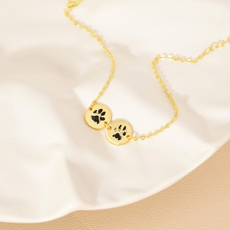 May include: A gold necklace with two circular pendants, each displaying a black paw print. The necklace is shown on a white, textured surface. The chain and pendants are delicate, showcasing the jewelry's design.