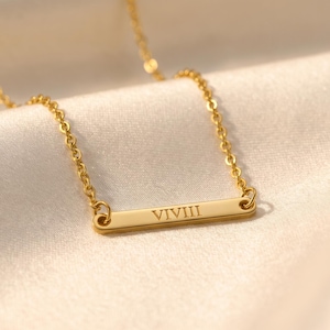May include: A gold-colored necklace with a rectangular pendant. The pendant has the Roman numeral "VIVIII" engraved on it. The necklace is displayed on a cream-colored, silky fabric.
