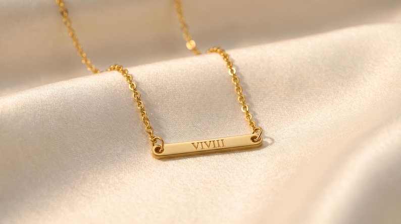 May include: A gold-colored necklace with a rectangular pendant. The pendant has the Roman numeral "VIVIII" engraved on it. The necklace is displayed on a cream-colored, silky fabric.