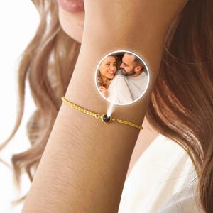 May include: A gold-colored bracelet with a small, dark-colored central stone. The bracelet is on a person's wrist, and a circular image of a couple is superimposed on the wrist. The bracelet has a delicate chain design.