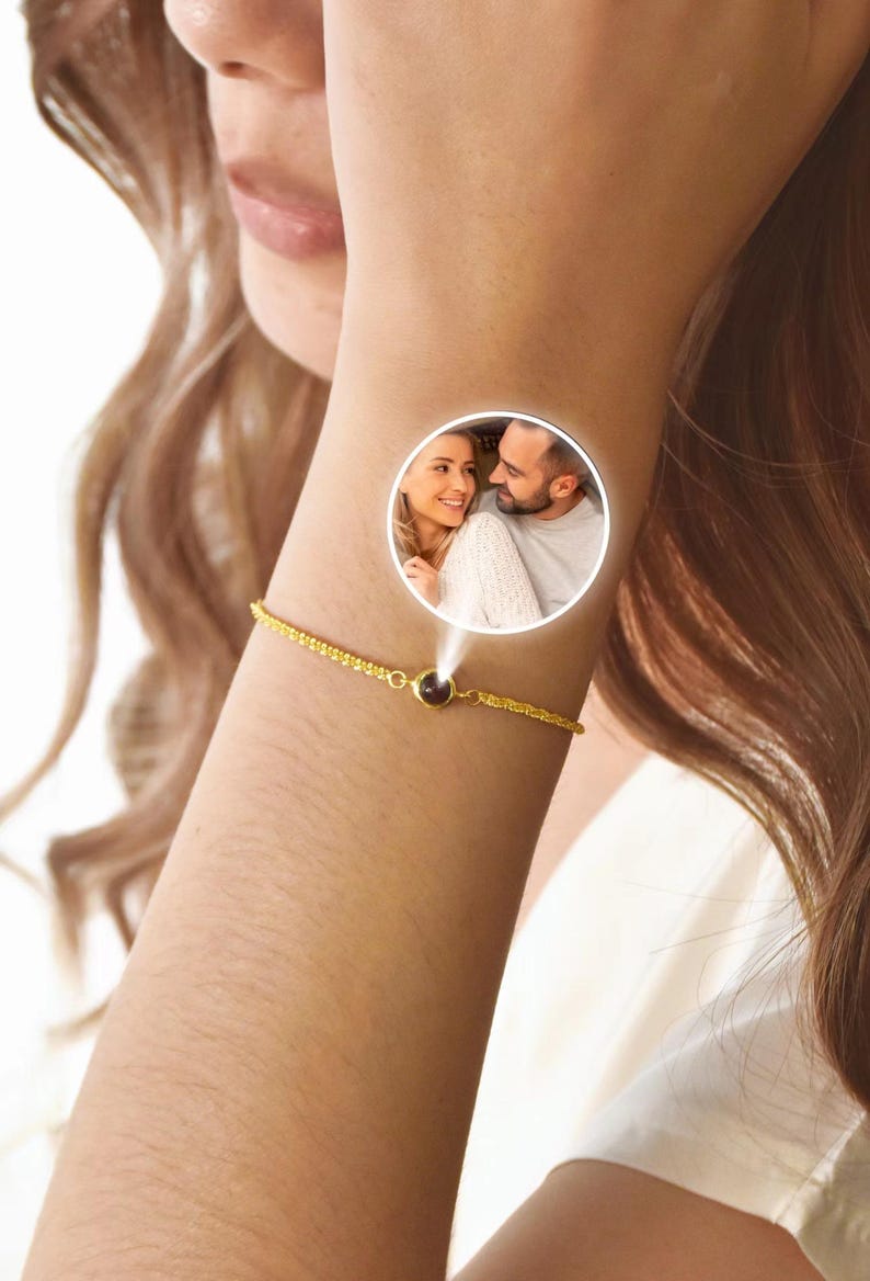 May include: A gold-colored bracelet with a small, dark-colored central stone. The bracelet is on a person's wrist, and a circular image of a couple is superimposed on the wrist. The bracelet has a delicate chain design.
