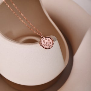 May include: A rose gold necklace with a paw print charm. The charm is round and has a raised paw print design. The necklace is on a white surface.