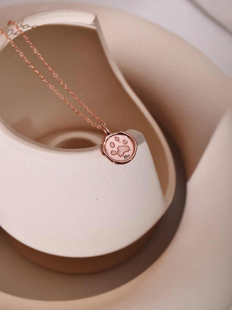 May include: A rose gold necklace with a paw print charm. The charm is round and has a raised paw print design. The necklace is on a white surface.