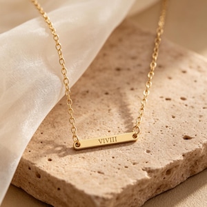 May include: A gold-colored necklace with a rectangular pendant. The pendant has the engraved text "VIVIII" and is suspended from a delicate chain. The necklace is displayed on a textured, light-colored surface.