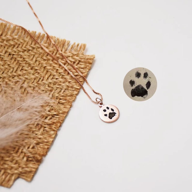 Op de afbeelding: Een ros&eacute;gouden ketting met een kleine, ronde hanger met een zwarte pootafdruk. De fijne ketting ligt op een getextureerd, bruin oppervlak. Een aparte pootafdruk is zichtbaar op een klein, rond, grijs oppervlak.
