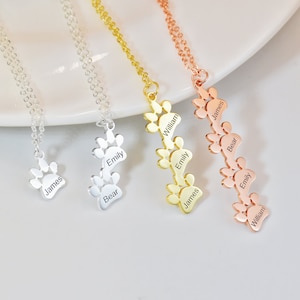 May include: Three silver, gold, and rose gold necklaces with paw print charms. Each charm has a name engraved on it: James, Bear, Emily, William. The necklaces are on a white surface.