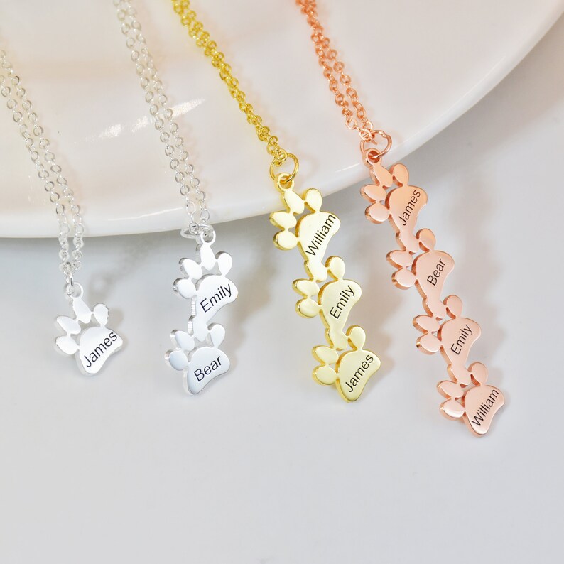May include: Three silver, gold, and rose gold necklaces with paw print charms. Each charm has a name engraved on it: James, Bear, Emily, William. The necklaces are on a white surface.