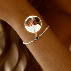 May include: A silver bracelet with a small, round, dark red gemstone. The bracelet is worn on a person's arm. A circular image of a couple kissing is superimposed on the arm.