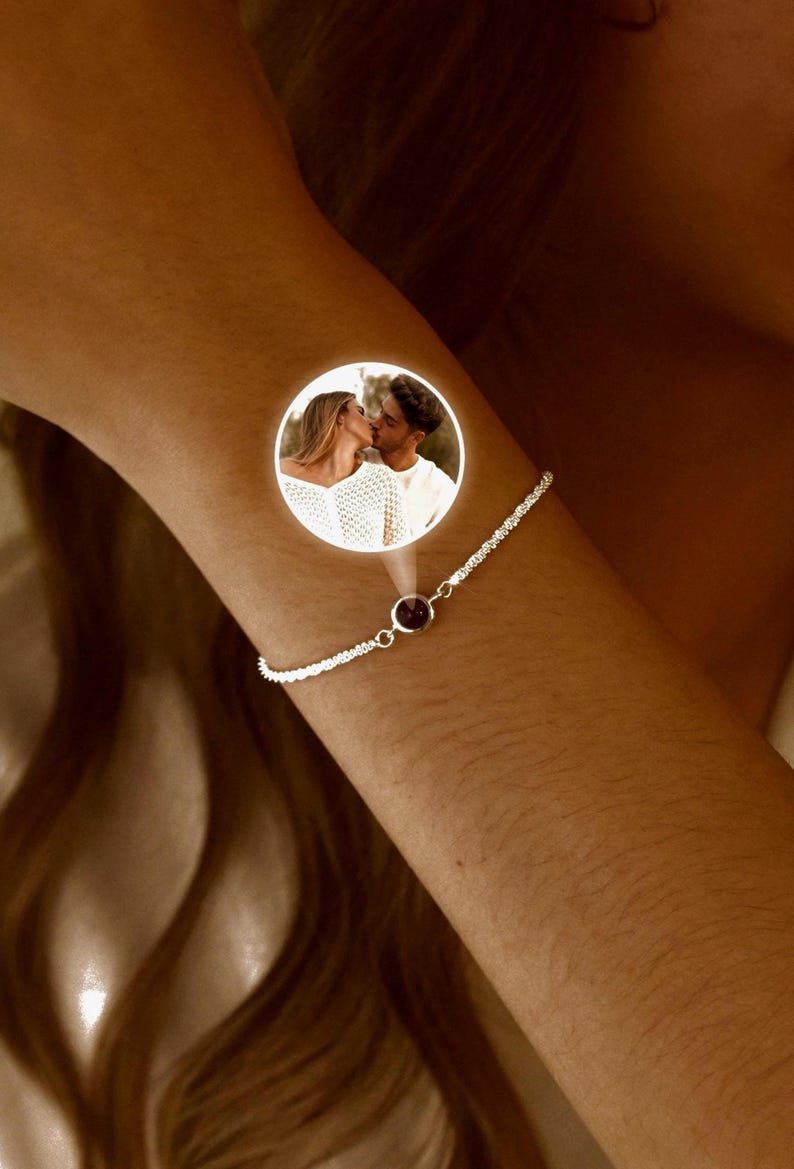 May include: A silver bracelet with a small, round, dark red gemstone. The bracelet is worn on a person's arm. A circular image of a couple kissing is superimposed on the arm.