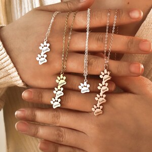 May include: Three silver necklaces with paw print charms. Each charm has a different name engraved on it: Emily, Bear, James, William. The necklaces are layered on top of each other.