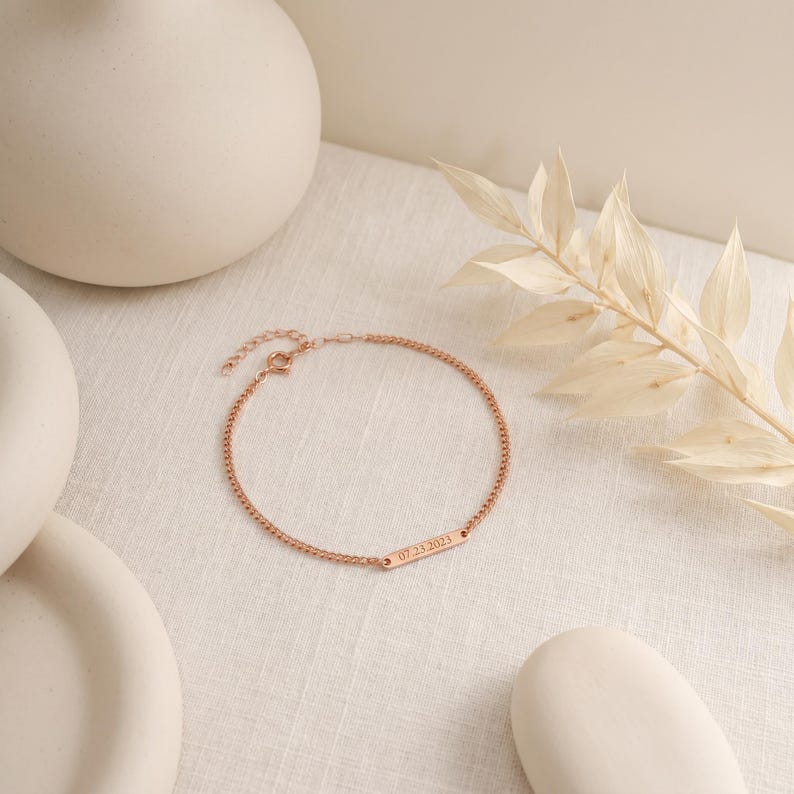 May include: A rose gold-toned chain bracelet featuring a small, rectangular metal bar engraved with the date 07.23.2023. The delicate jewelry piece includes a thin link chain and a circular clasp mechanism for secure fastening around the wrist.