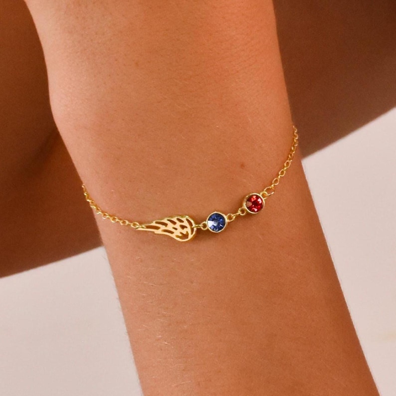 Pode incluir: Uma pulseira dourada com um delicado pingente de asa e dois detalhes de pedras preciosas. A pulseira de corrente apresenta uma gema azul e uma vermelha. A pulseira &eacute; usada em um bra&ccedil;o.