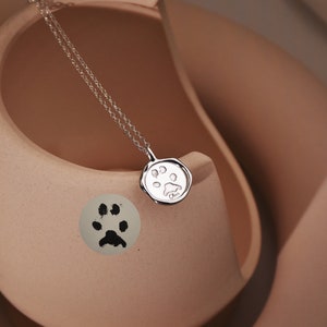 May include: A silver necklace with a paw print charm. The necklace is displayed on a light brown ceramic surface.