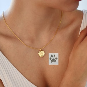 May include: A gold chain necklace with a small round pendant featuring a paw print. The pendant is on a woman's chest. A paw print is also visible on a white background.