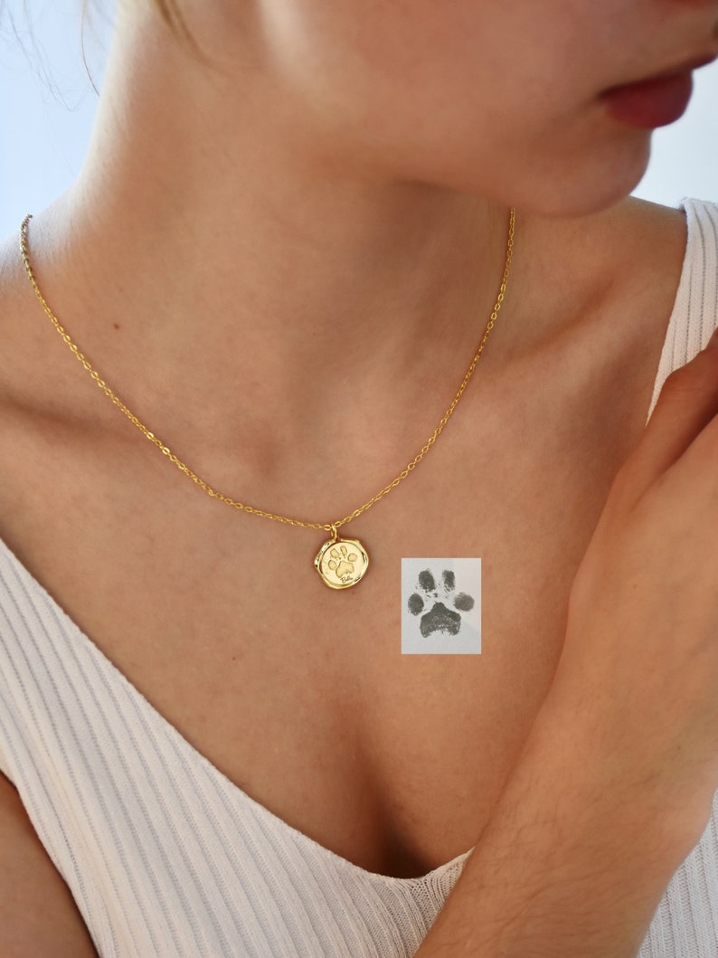May include: A gold chain necklace with a small round pendant featuring a paw print. The pendant is on a woman's chest. A paw print is also visible on a white background.