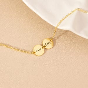 May include: A gold-coloured necklace featuring two circular pendants. Each pendant is inscribed with a name in cursive script: "Findus" and "Gipsy". The necklace chain is delicate, set against a soft, neutral background.