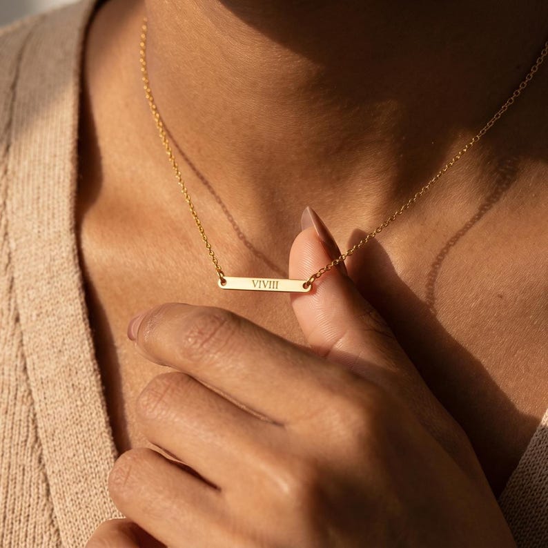 May include: A gold-colored bar necklace with the Roman numeral "VIIII" engraved on the bar. The necklace is on a delicate chain and is being held by a person's hand. The background is a neutral beige.