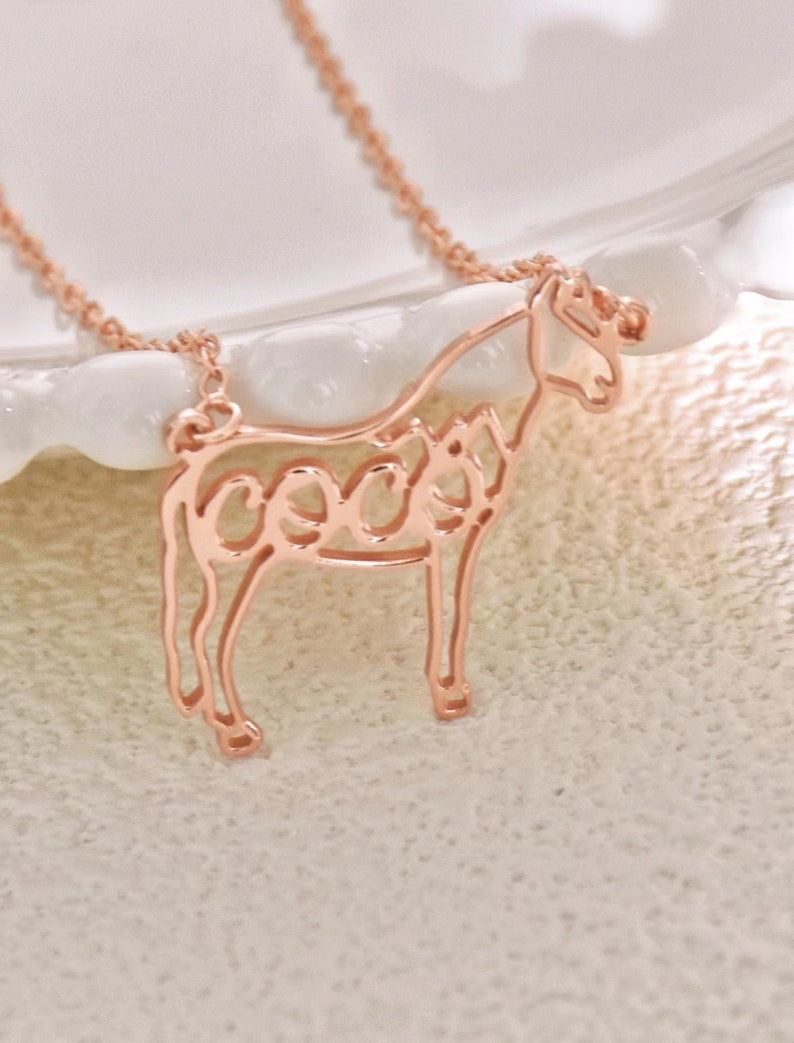 May include: A rose gold necklace with a horse pendant. The horse is outlined and the word "coco" is spelled out within the outline.
