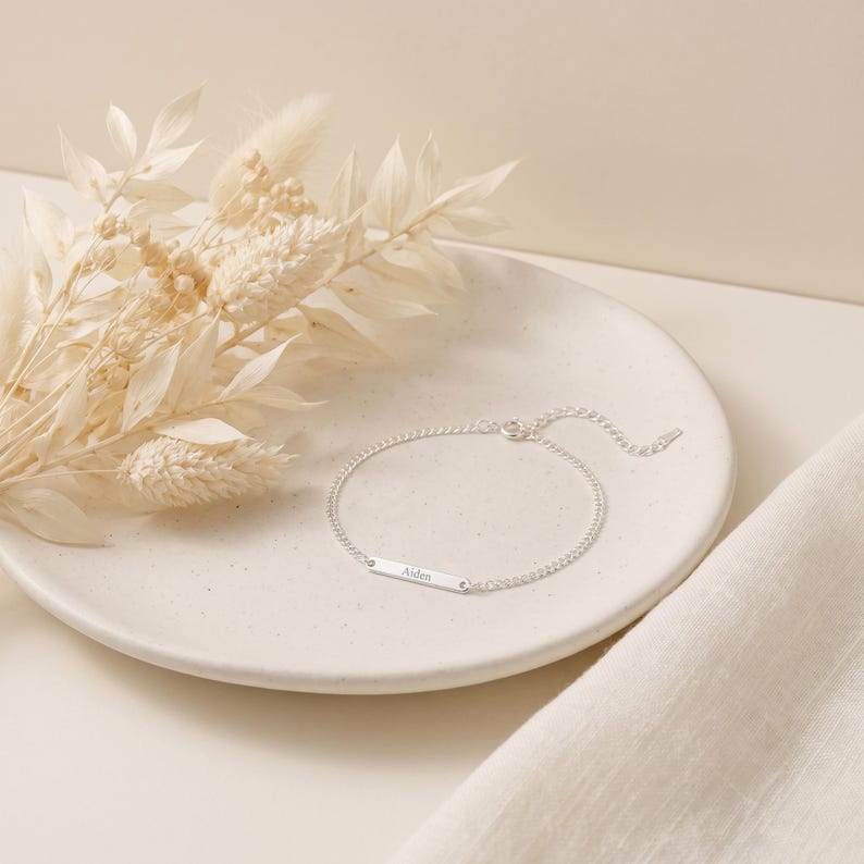 May include: A silver-toned chain bracelet featuring a thin, rectangular metal bar engraved with the name Aiden. The bracelet includes a delicate link chain and a small circular clasp, presenting a clean and simple design for everyday wear.