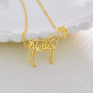 May include: Gold necklace with a delicate horse silhouette pendant. The horse's body is formed by the name "Heller" in a cursive font.