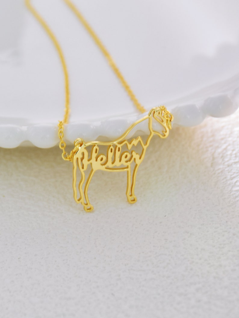 May include: Gold necklace with a delicate horse silhouette pendant. The horse's body is formed by the name "Heller" in a cursive font.