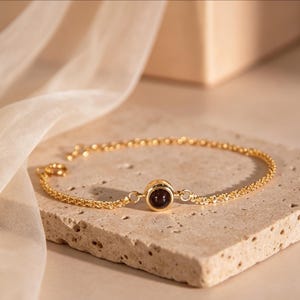May include: A gold-colored bracelet with a dark red gemstone in the center. The bracelet is made of a delicate chain and is displayed on a textured, light-colored surface. The background features a soft, draped fabric.