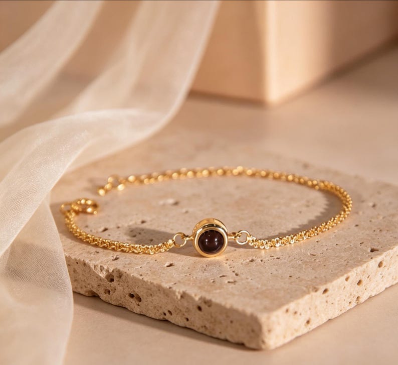 May include: A gold-colored bracelet with a dark red gemstone in the center. The bracelet is made of a delicate chain and is displayed on a textured, light-colored surface. The background features a soft, draped fabric.