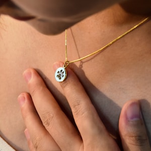 Op de afbeelding: Een gouden ketting met een kleine, ovale hanger. De hanger heeft een witte achtergrond met een zwart pootafdrukontwerp. De ketting wordt om de nek gedragen.