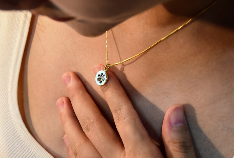 Op de afbeelding: Een gouden ketting met een kleine, ovale hanger. De hanger heeft een witte achtergrond met een zwart pootafdrukontwerp. De ketting wordt om de nek gedragen.