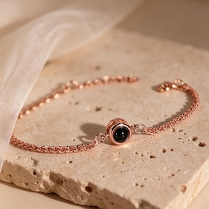 May include: Rose gold bracelet with a delicate chain and a central round charm. The charm features a black stone, adding a touch of contrast to the warm metallic hue. The bracelet is displayed on a textured, light-colored surface.