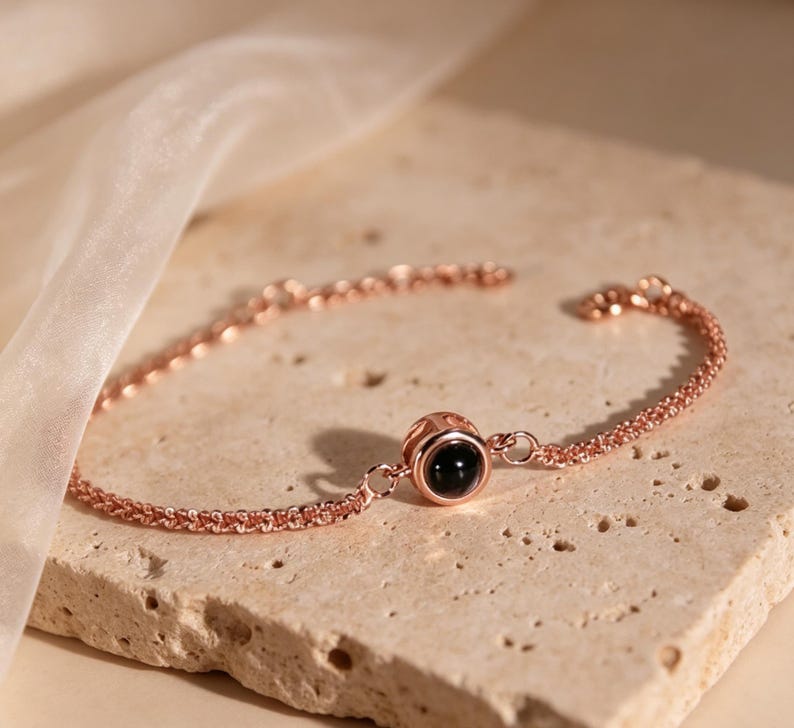 May include: Rose gold bracelet with a delicate chain and a central round charm. The charm features a black stone, adding a touch of contrast to the warm metallic hue. The bracelet is displayed on a textured, light-colored surface.
