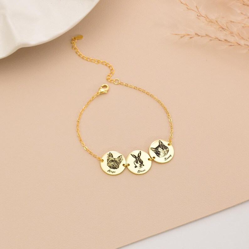 May include: A gold-coloured bracelet with three circular pendants, each engraved with a pet portrait. The pendants show a dog, a rabbit, and a cat, with names beneath. The bracelet is displayed on a pale pink surface.