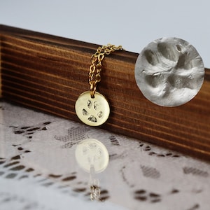 Op de afbeelding: Een goudkleurige ketting met een hanger in de vorm van een pootafdruk. De ronde hanger hangt aan een fijne ketting. Een close-up van de pootafdruk is te zien. De ketting ligt op een houten oppervlak.