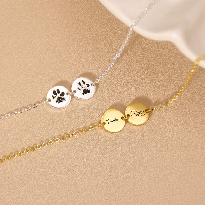May include: Two necklaces are shown, one silver and one gold. The silver necklace has two circular pendants with paw prints. The gold necklace features two pendants with the names "Findus" and "Gipsy" engraved on them.